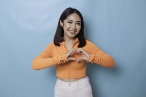 séduisante jeune femme asiatique se sent heureuse et romantique formes coeur geste exprime des sentiments tendres porte un haut orange décontracté sur fond bleu. concept d'affection et de soins des personnes photo