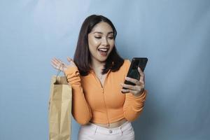 portrait asiatique heureuse belle jeune femme debout excitée tenant un sac à provisions en ligne et un smartphone à portée de main, prise de vue en studio isolée sur fond bleu photo