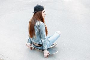 jolie femme dans une casquette de baseball et des vêtements en denim assis sur une planche à roulettes sur le fond de l'asphalte photo