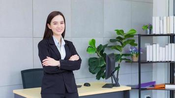 jeune femme d'affaires professionnelle asiatique qui a les cheveux longs porte un costume formel noir avec une chemise bleue pendant qu'elle croise les bras et sourit joyeusement au bureau. photo