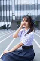 belle lycéenne asiatique en uniforme scolaire avec des sourires confiants pendant qu'elle regarde la caméra avec bonheur avec le bâtiment en arrière-plan. photo