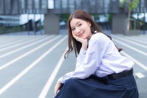 une lycéenne asiatique en uniforme scolaire avec des bretelles sur les dents est assise et sourit avec confiance pendant qu'elle regarde la caméra avec bonheur avec le bâtiment en arrière-plan photo