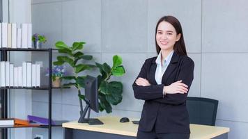 jeune femme d'affaires professionnelle asiatique qui a les cheveux longs porte un costume formel noir avec une chemise bleue pendant qu'elle croise les bras et sourit joyeusement au bureau. photo
