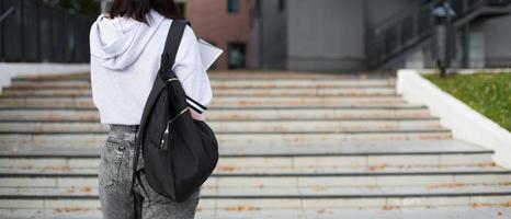 une étudiante avec un sac à dos noir et un cahier dans les mains se tient sur les marches devant l'institut. l'enseignement supérieur, la rentrée, la rentrée. fond photo