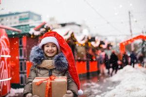 portrait d'une fille joyeuse en bonnet de noel avec boîte-cadeau pour noël dans la rue de la ville en hiver avec de la neige sur le marché festif avec des décorations et des guirlandes lumineuses. vêtements chauds, écharpe tricotée et fourrure. nouvel An photo