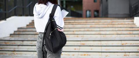 une étudiante avec un sac à dos noir et un cahier dans les mains se tient sur les marches devant l'université. l'enseignement supérieur, la rentrée, la rentrée. fond