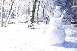 gros bonhomme de neige dans la cour après une chute de neige en hiver. nez-carotte, seau sur la tête, gants sur les mains, yeux. voitures dans la neige le long de la route. activités extérieures. copie espace photo