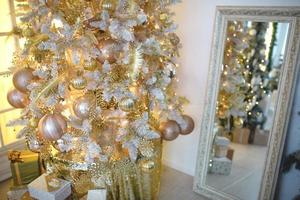 un arbre de noël avec une décoration dorée se reflète dans le miroir. nouvel an, décoration de la maison. grande fenêtre avec lumière jaune chaude, confort dans le salon photo