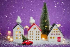 décor de noël - une voiture rétro rouge sur la neige passe devant des maisons avec des guirlandes lumineuses dans un arbre de noël bokeh avec des coffrets cadeaux sur le toit. jouet sur fond violet. carte de voeux de nouvel an. maison confortable photo