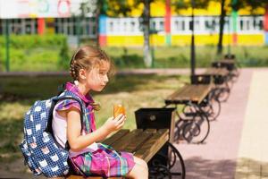 fille avec un sac à dos assis sur un banc et mangeant une tarte près de l'école. une collation rapide avec un petit pain, des aliments malsains, un déjeuner à la maison. retour à l'école. éducation, classes d'école primaire, 1er septembre photo