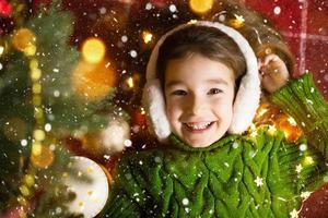 une fille heureuse dans des écouteurs en fourrure se trouve près de l'arbre de noël dans des guirlandes et des lumières dans un pull chaud sur une couverture confortable. noël et nouvel an, veille de vacances, ambiance magique photo