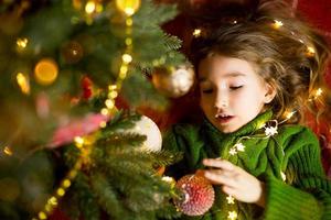 une fille aux cheveux longs et aux guirlandes se trouve sur un plaid rouge sous un sapin de noël avec des jouets dans un pull tricoté chaud. noël, nouvel an, émotions des enfants, joie, attente d'un miracle et cadeaux photo