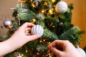 mains de femme décorant le sapin de noël. sapin de noël avec décorations blanches et argentées, jouet d'ornements et balle. design d'intérieur de style classique moderne. réveillon de noël à la maison, temps de fête. photo