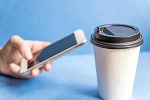 gobelet jetable en papier kraft blanc pour café avec couvercle en plastique noir et téléphone portable blanc dans une main féminine sur fond bleu. photo