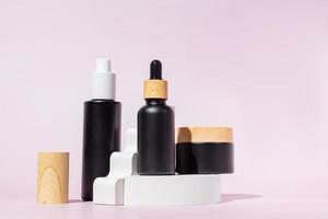 trois bouteilles en verre noir aux formes géométriques tendance sur fond rose. socle et échelle blancs. produits de soins de la peau, cosmétiques naturels. concept de beauté pour les soins du visage photo