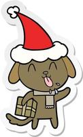 dessin animé mignon autocollant d'un chien avec un cadeau de noël portant un bonnet de noel vecteur