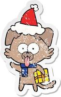 autocollant en détresse dessin animé d'un chien avec un cadeau de noël portant un bonnet de noel vecteur