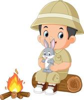 un aventureux garçon est assis sur une déchue arbre et détient une lapin tandis que éclairage une feu vecteur