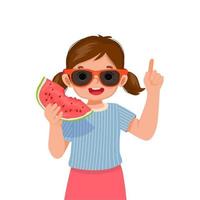 heureuse petite fille mignonne avec des lunettes de soleil mangeant de la pastèque pointant le doigt vers le haut le jour ensoleillé de l'heure d'été vecteur
