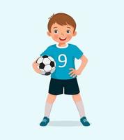 portrait d'un mignon petit garçon en vêtements de sport tenant un ballon de football posant avec la main sur la taille vecteur