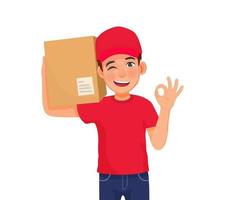 jovem entregador ou serviço de correio com uniforme de boné vermelho segurando o pacote de caixa no ombro, mostrando o gesto de sinal okey vetor