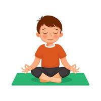 menino bonitinho fazendo exercícios de ginástica praticando meditação ioga sentado em pose de lótus em um tapete verde interior em casa vetor