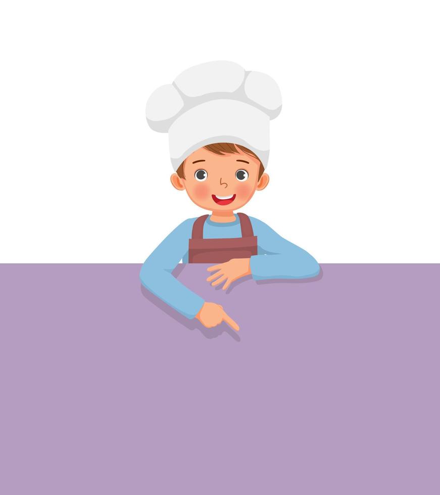 ragazzino carino con cappello da cuoco e grembiule in piedi dietro una tavola vuota che punta il dito verso un banner vuoto per lo spazio della copia vettore