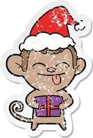 divertente adesivo angosciato cartone animato di una scimmia con regalo di Natale che indossa il cappello di Babbo Natale vettore