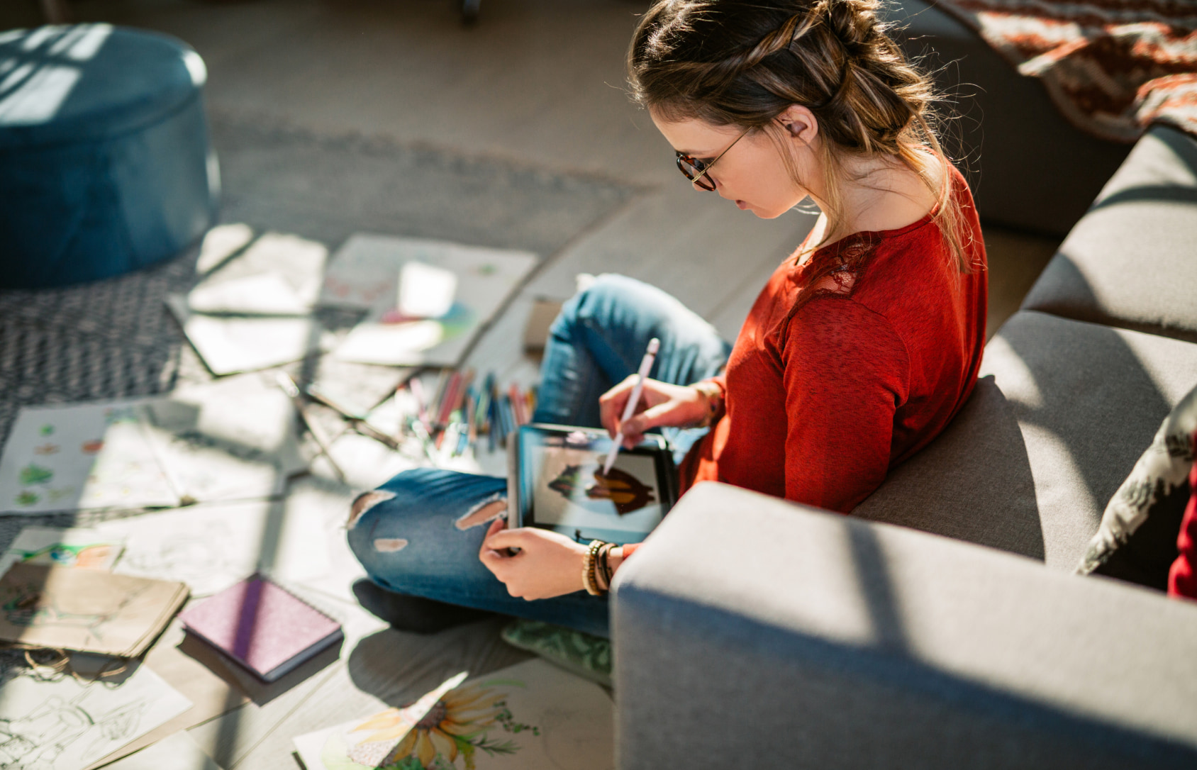 Young woman wearing glasses, sitting in front of a couch, drawing on tablet.